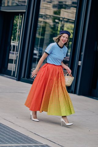 Chloe King at New York Fashion Week in a Christopher John Rogers orange and yellow polka dot skirt, a blue top, blue hat, and white heels