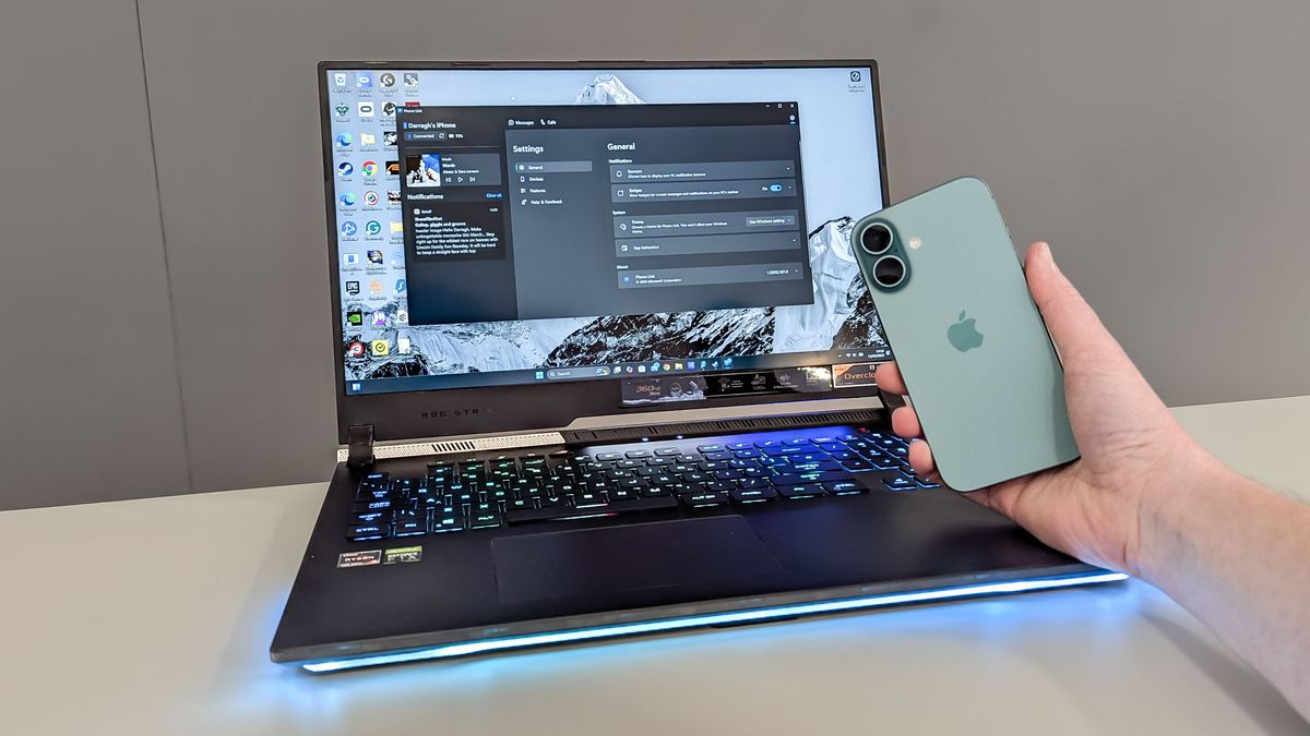 iPhone 16 in hand being held in front of Asus ROG Strix 17 on table showing Phone Link app