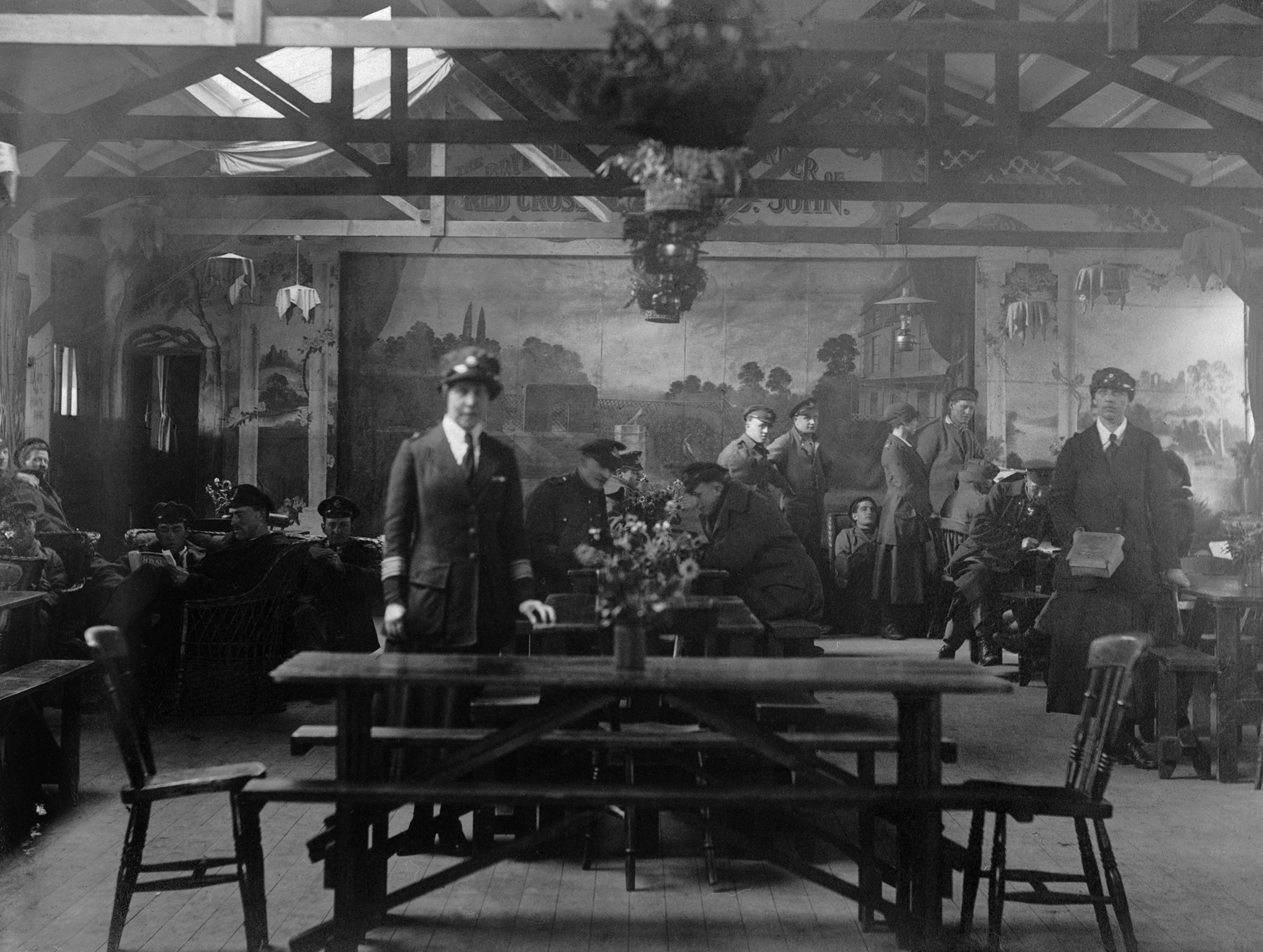 Women At Work During The First World War: Voluntary Aid Detachment, A general view of the interior of the British Red Cross Society and OSJJ Recreation Hut at Wimereux. In the foreground can be seen VAD Commandant Miss Barber, standing at a table. In the background, chatting to some of the soldiers are Miss Scott (background right) and Miss Dixon (right). A large mural can be seen on the back wall, circa 1916. (Photo by Olive Edis / Imperial War Museums via Getty Images)