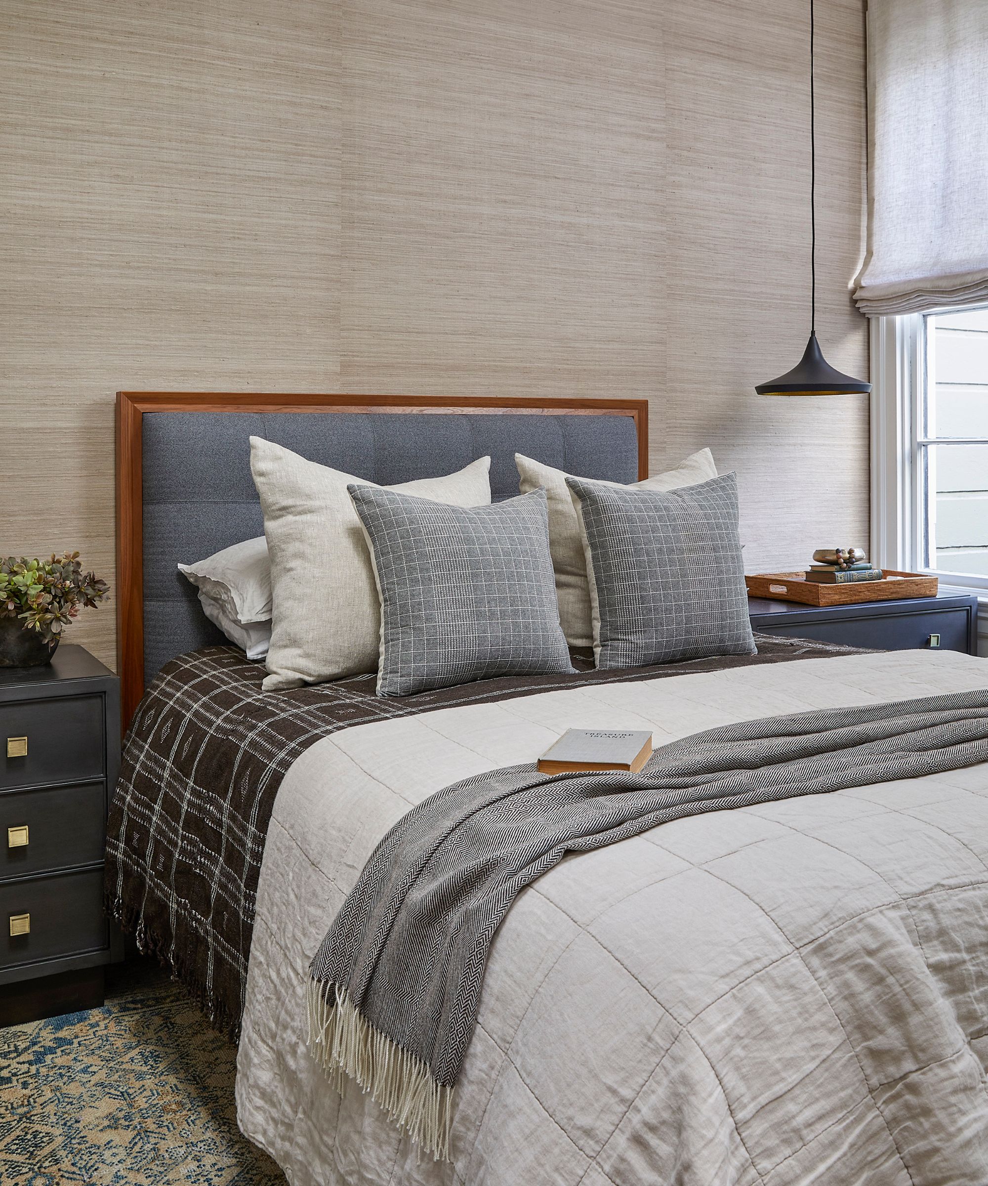 Neutral bedroom with grasscloth wallpaper, a gray upholstered headboard, and dark gray nightstands with brass hardware