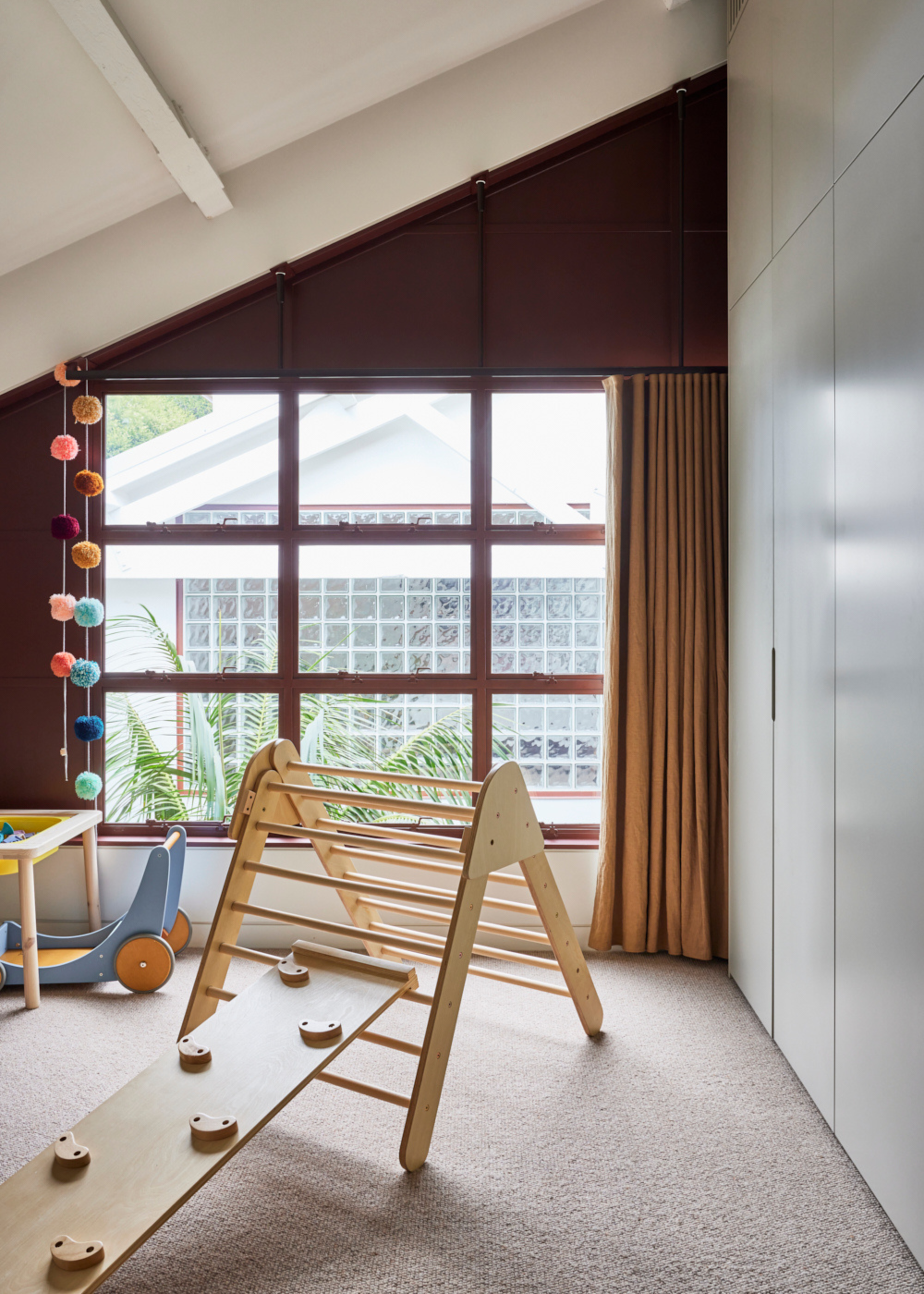 A white kids room with a brown wall with windows, a wood climbing toy, patterned lanterns, and brown window treatments