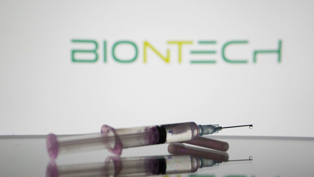 A medical syringe sits on a reflective surface, in front of the BioNTech logo (the word &amp;#039;BioNTech&amp;#039; stylised in shades of green) displayed on a wall