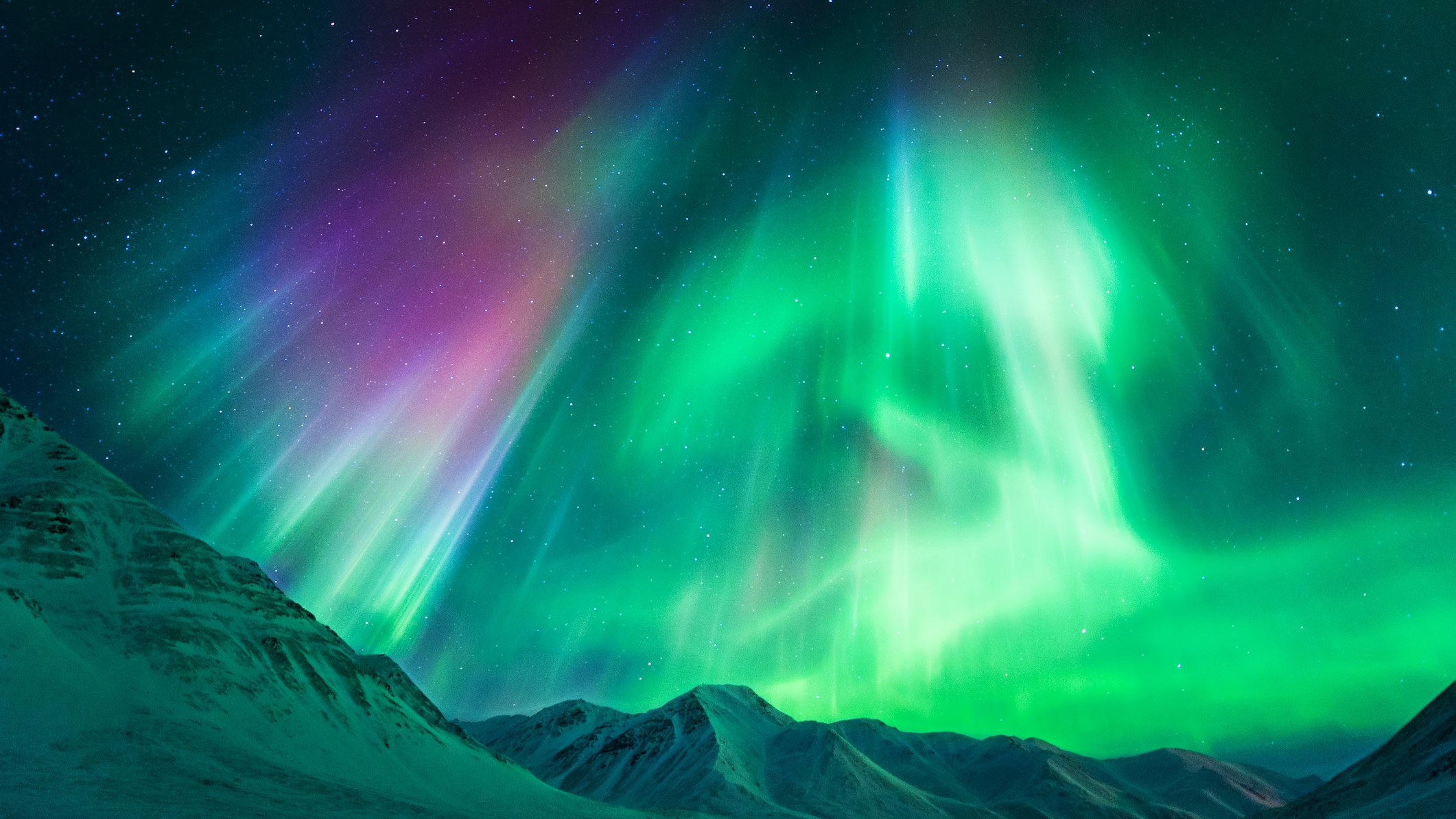 Ribbons of green and purple light appear in the sky above snow capped mountain peaks.