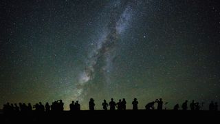 Silhouettes of people are seen standing on the ground, looking up at a starry sky and the center of the Milky Way.