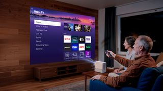 A middle-aged couple are very pleased with the Aurzen Roko TV Smart Projector, which is projecting the Roku TV interface onto a screen in their living room