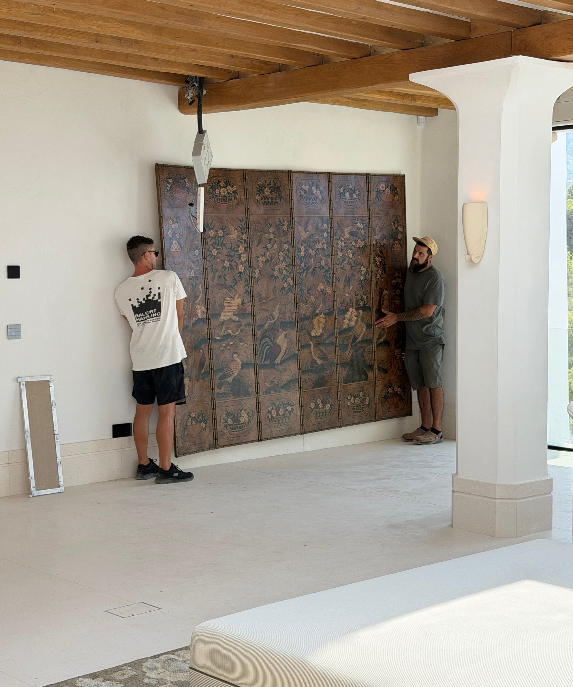 Two men holding a large folding screen against a white wall, whie concrete floor and pillar in foreground