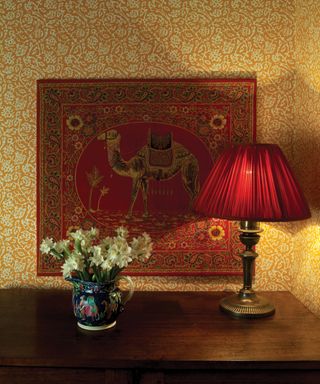 Hallway decorated with floral, yellow wallpaper, dark wood table with flowers and petite red lampshade, red wall art with intricate detailing of flowers and a camel