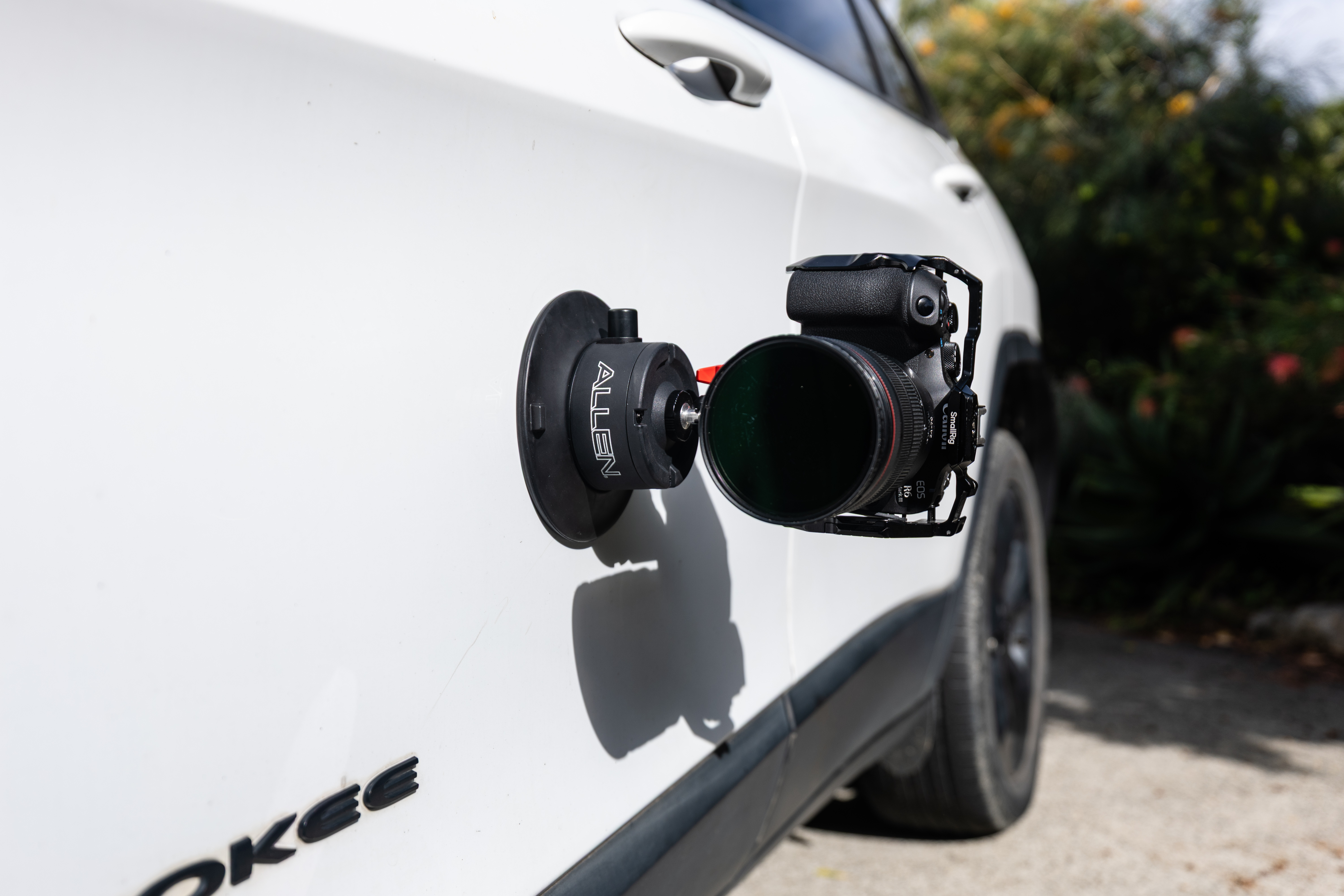 An Allen Smart Suction Snap Camera Mount is fixed vertically to the side of a white Jeep, securely holding a Canon mirrorless camera equipped with a professional lens.