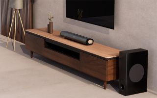 Lifestyle shot of the Creative Stage Pro soundbar on a wooden unit below a TV. The carpet and walls are a kind of oatmeal color