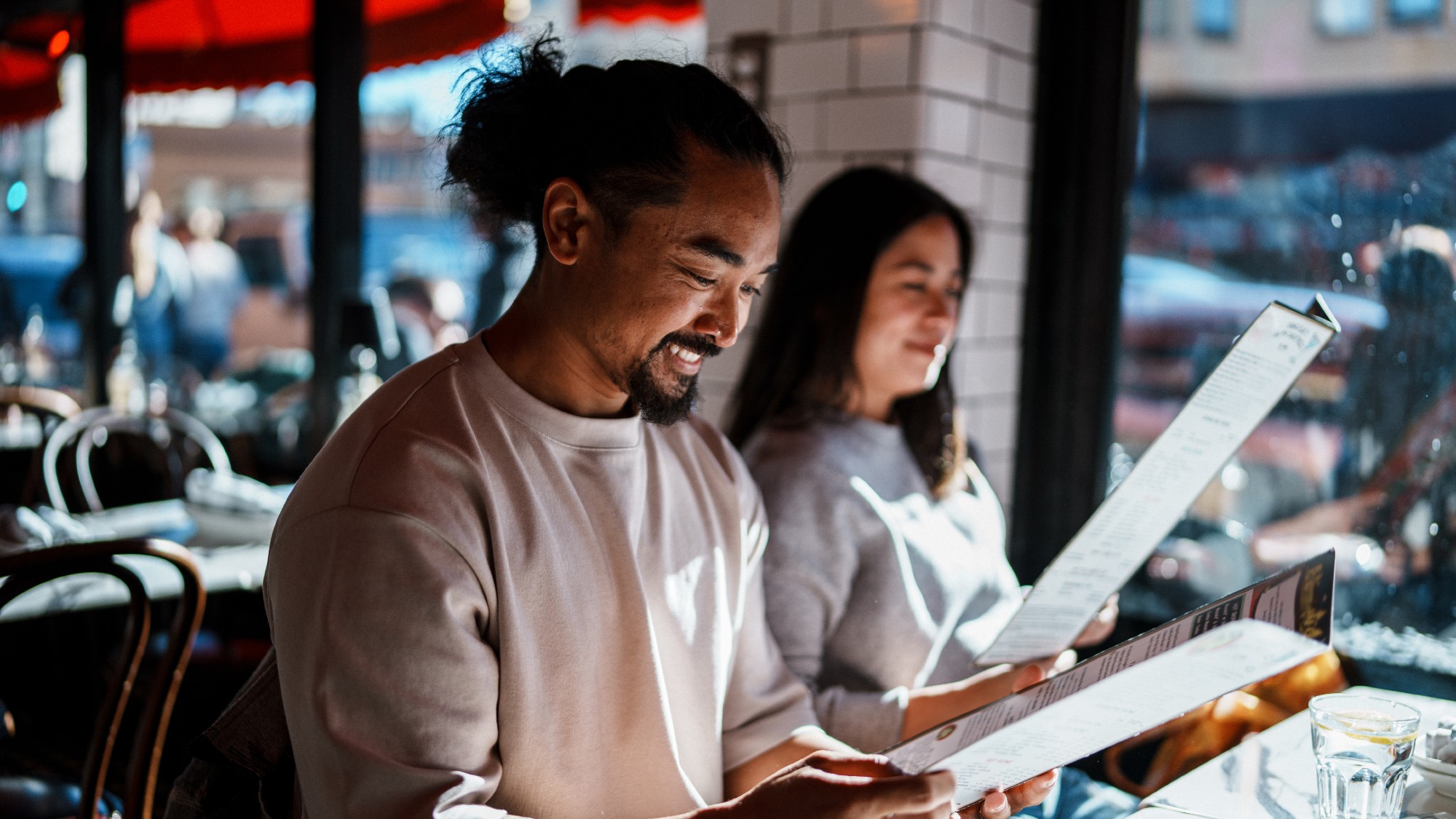 a couple sits at a counter of a restaurant. they are both looking at the menu. the man has a goatee and hair in a ponytail. the woman has shoulder length hair