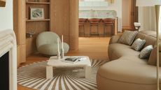 Image of a modern living room that opens to a kitchen. The living room has wooden floors and walls, a beige leather bean-shaped sofa, a spiral rug, and a white marble coffee table.