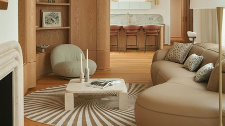 Image of a modern living room that opens to a kitchen. The living room has wooden floors and walls, a beige leather bean-shaped sofa, a spiral rug, and a white marble coffee table.