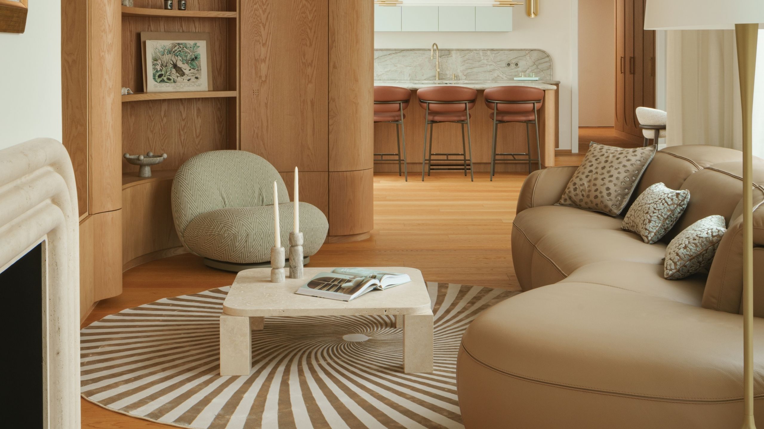 Image of a modern living room that opens to a kitchen. The living room has wooden floors and walls, a beige leather bean-shaped sofa, a spiral rug, and a white marble coffee table.