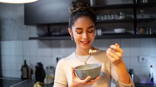 A girl with brown hair tied up in a bun eats dinner from a grey ceramic bowl in her kitchen at night