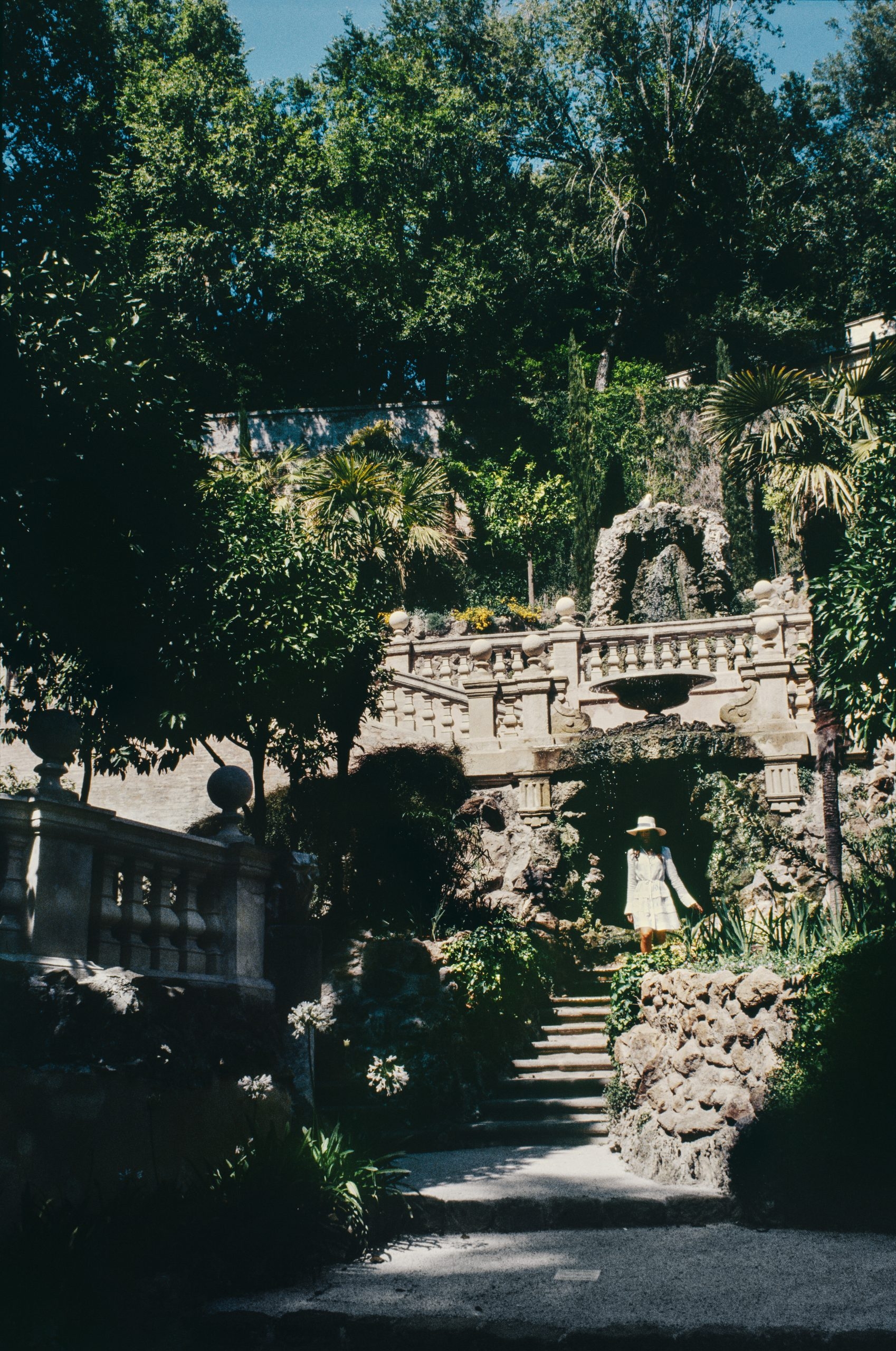 A lush, multi-level garden with marble architecture, textural grottos, colorful flowers, and blue skies.