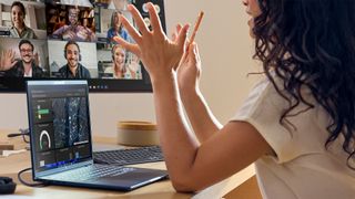 A woman with dark hair and a white top is videoconferencing with multiple people and referring to her HP Elitebook laptop