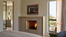 A white room with a fireplace on the wall and a window beside the fireplace. The window has layered curtains framing it and there is a doorway to a spacious bathroom in the left frame.