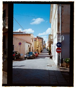 A film scan of a photo of a street in Sardinia with old buildings, parked cars and a blue sky