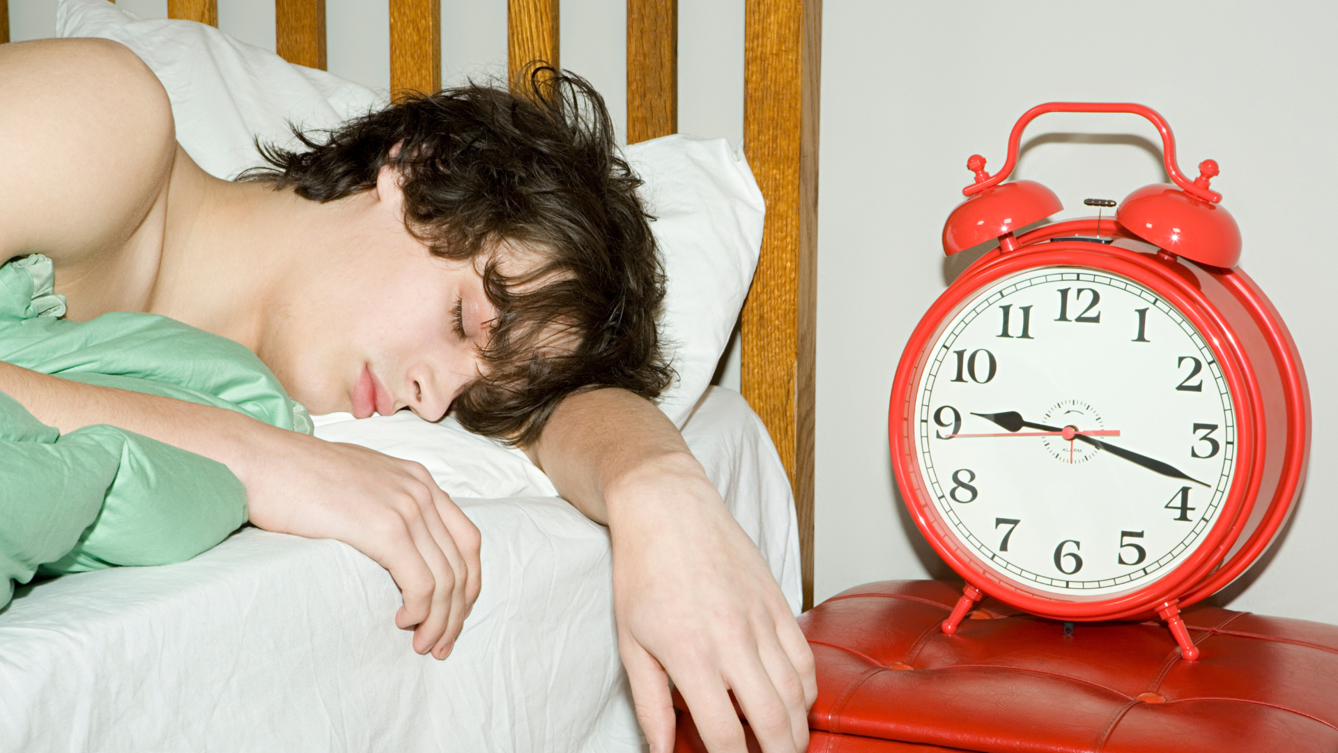 A teenage boy lies asleep in bed. The clock on his bedside table shows that it is around 9.20am.