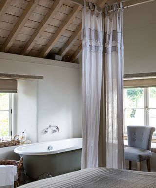 Rustic, country bedroom with exposed beams on wooden vaulted ceiling, wooden poster bed with soft sheer curtains, bathtub beside window
