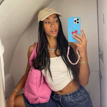 Dutch fashion influencer Amaka Hamelijnck taking a selfie on an airplne wearing a white tank, baseball hat, denim shorts, and pink tote bag