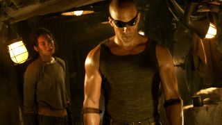 Still from the movie series The Chronicles of Riddick. Here we see Riddick (bald man with dark goggles) standing in a dark prison. Behind him in a woman in similar clothes.