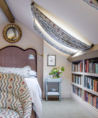 bedroom with cream walls in attic room with bookshelves and blue scallop bedside table and bedend armchair in rose fabric