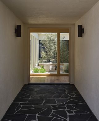 Image of an entryway hallway that has white walls and fractured slate flooring. The hall opens up into a wooden room that has a window view of an atrium.