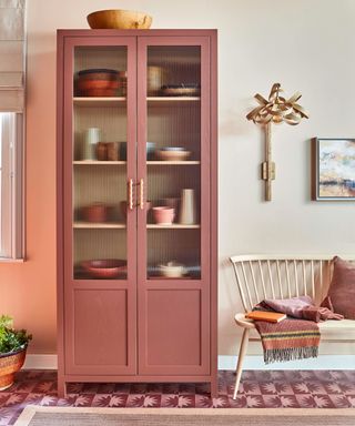 A kitchen with beige walls, a muted red cabinet with bowls on the shelves, a pale wooden chair with a striped red throw and books on it, and red and pink patterned carpet