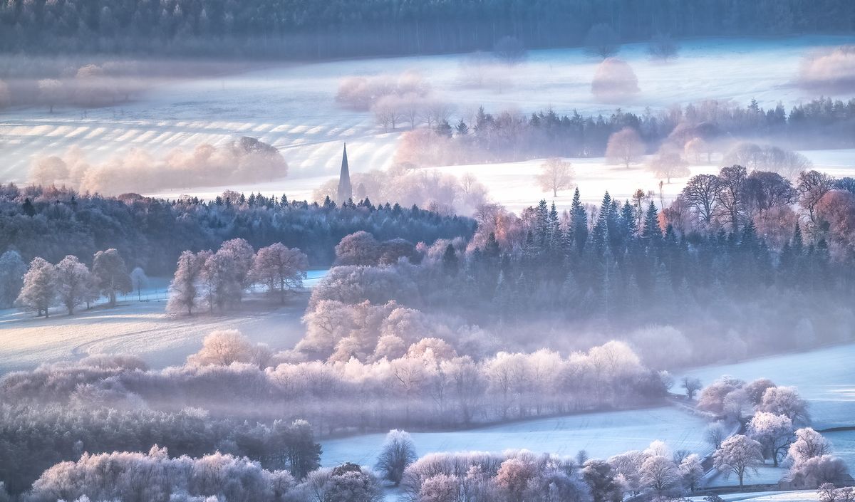 Frosty and misty landscape of the Peak District, with a church spire in the far distance surrounded by layers of open fields and forest