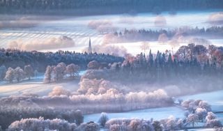 Frosty and misty landscape of the Peak District, with a church spire in the far distance surrounded by layers of open fields and forest