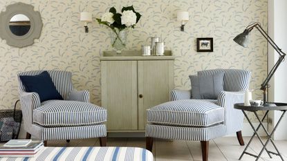 A bright and airy living room with patterned cream and blue wallpaper, two striped blue and white upholstered chairs, a pale green fluted cabinet with flowers on top, and a striped ottoman with a stack of books in the foreground.