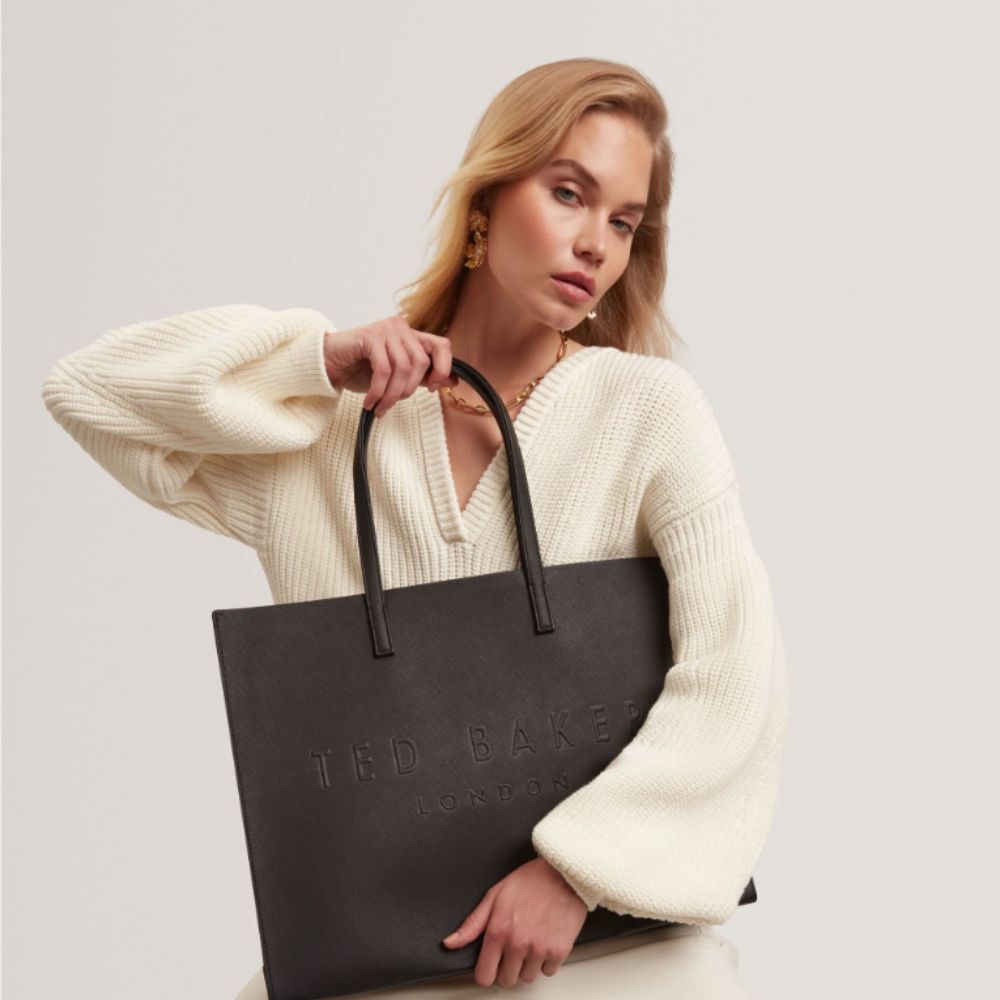 Young blonde woman in Ted Baker white jumper holding Ted Baker stone black bag.