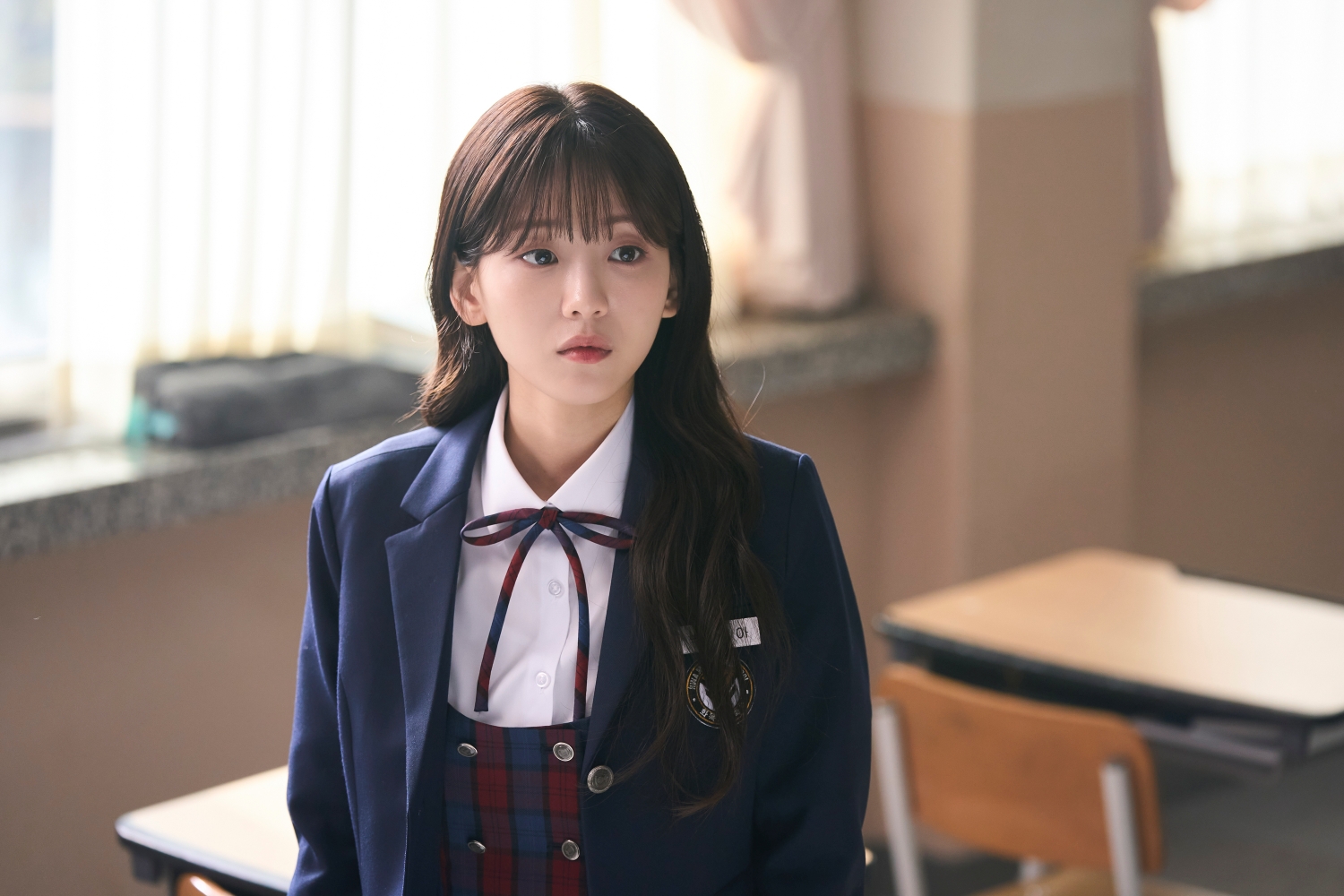 Cho Yi-hyun in the Prime Video K-drama 'Head Over Heels.' wearing a blazer and a ribbon around her neck sititng on a school desk