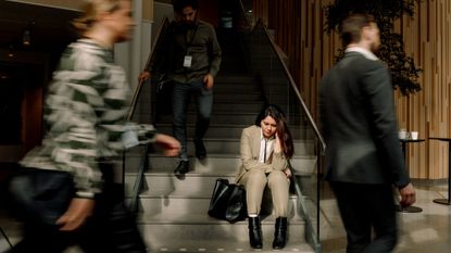 Frustrated woman in a business suit sitting on stairs while other workers walk around her in a blur 