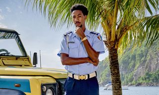 Officer Sebastian Rose (Shaquille Ali-Yebuah) stands next to the yellow police jeep in his uniform, with a palm tree behind him and a view of a coastal bay in the distance. He is resting his chin on his left hand and looking thoughtful.