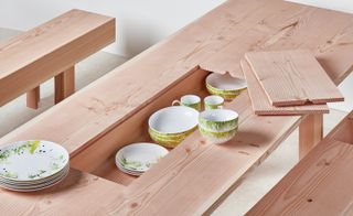 ’Planks’ collection by Max Lamb, natural wood table and seating benches, middle section of table open with white patterned teacups, bowls and small plates inside, six dinner plates stacked on the table top, two wood planks from the inner section placed on the table top, white floor and walls