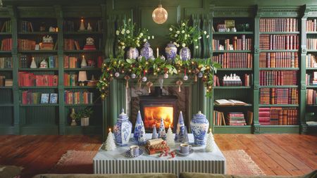 A living room with green built-in shelving, filled with books and Christmas decor, surrounding a lit fireplace with a garland hanging over the mantel 