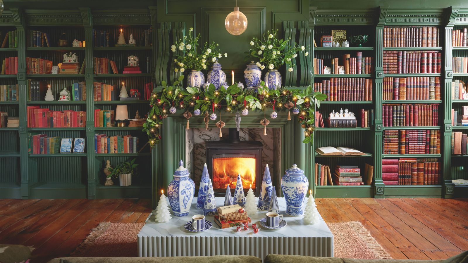 A living room with green built-in shelving, filled with books and Christmas decor, surrounding a lit fireplace with a garland hanging over the mantel 