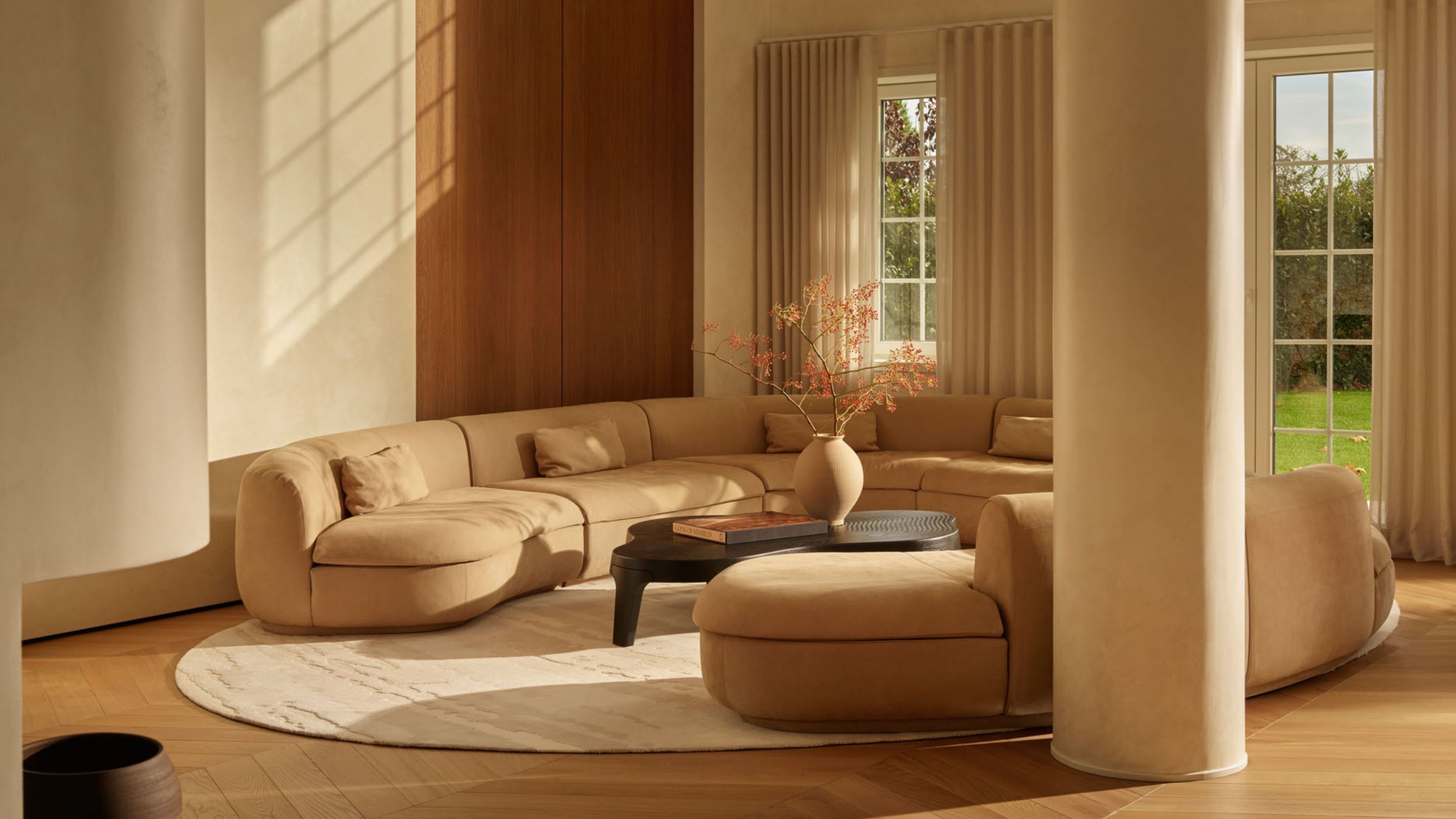 Image of a spacious minimalist living room with two large curved beige sofas facing each other on top of an oval white rug.