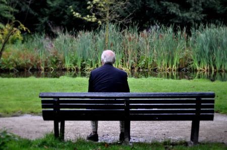 An older man sits alone on a bench by a lake abroad.