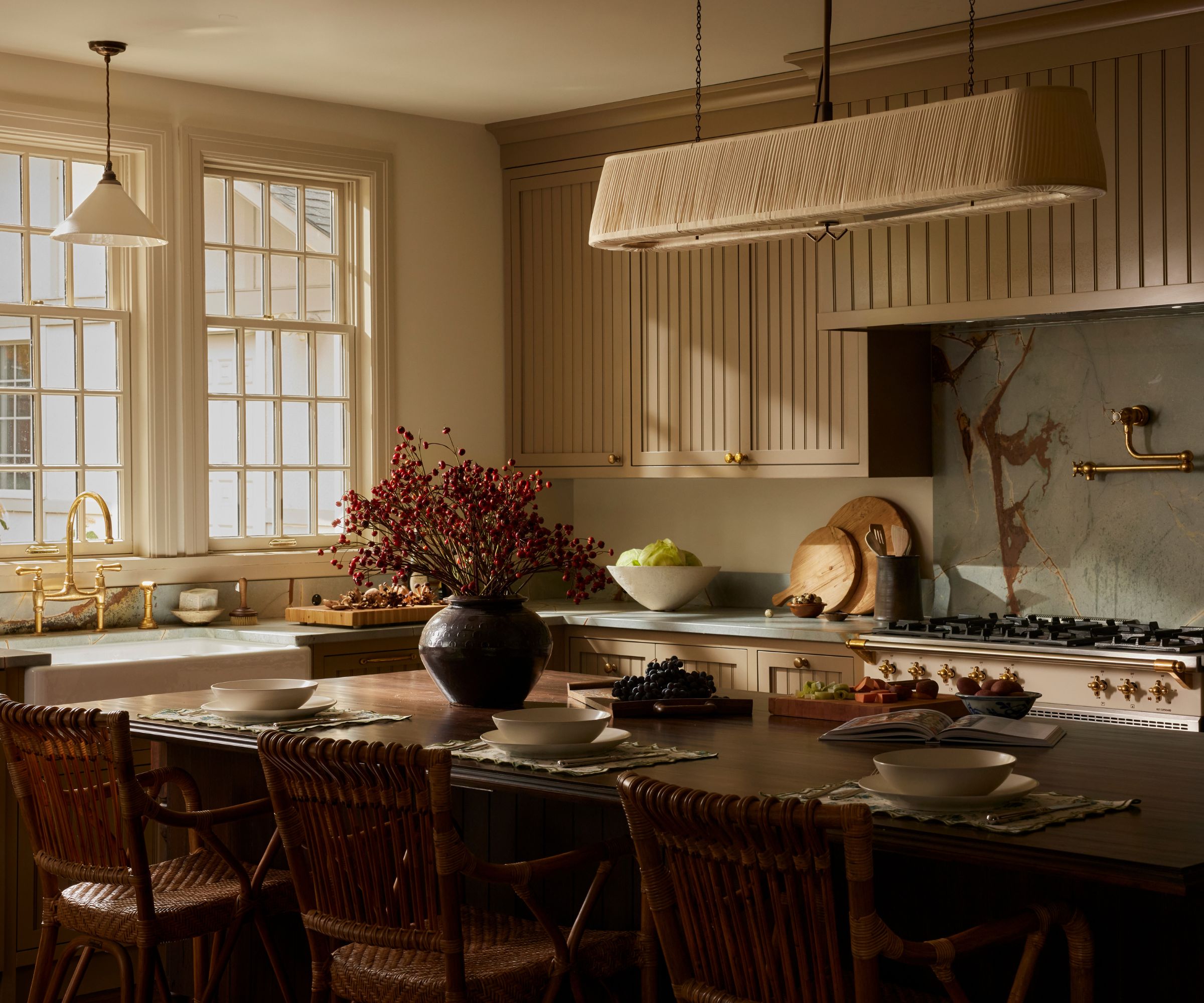 Cozy kitchen, large dark wood dining table in center, decorated with vase of flowers and ableware, slatted wooode kitchen cabinets, stove and windows, sink in background