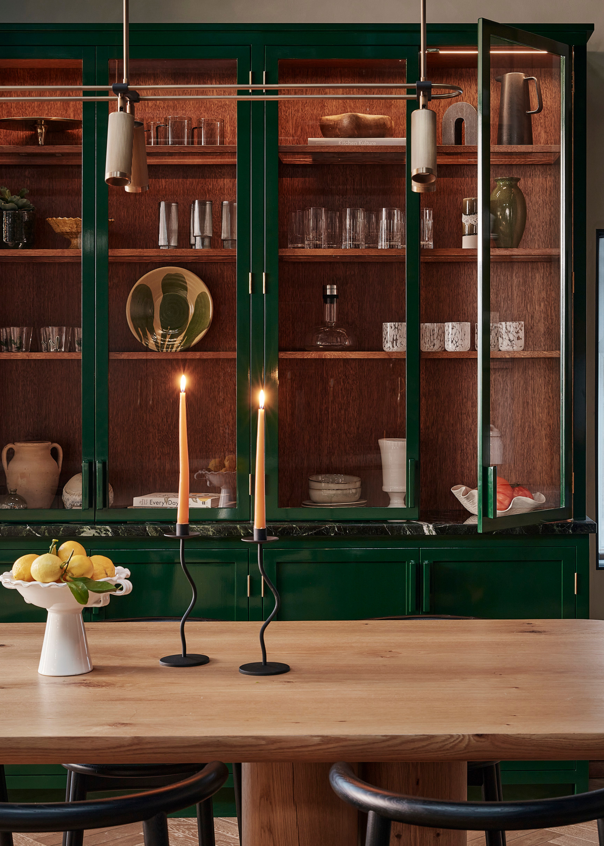 A modern kitchen with green gloss, glass-fronted kitchen cabinets filled with decorative glassware and tableware in front of a natural wood table with thick, circular legs with a fruit bowl and two lit taper candles on top with curved black dining chairs