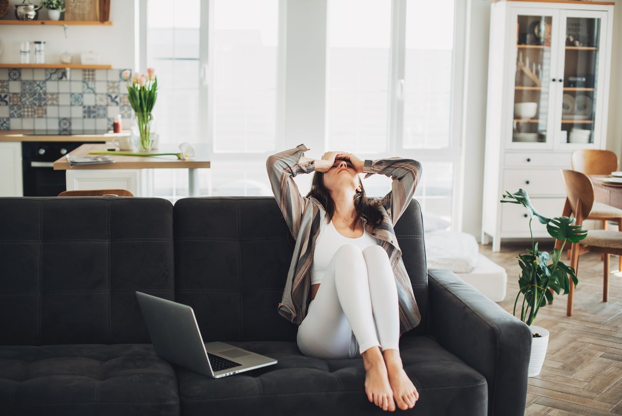 A young woman sits on her couch with her hands over her eyes in a gesture of regret.