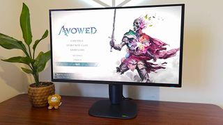 Alienware AW2725Q monitor on woodgrain desk next to plant with Avowed main menu on screen