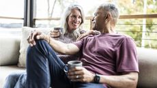 An older couple sit on their sofa with coffee cups and smile at each other.