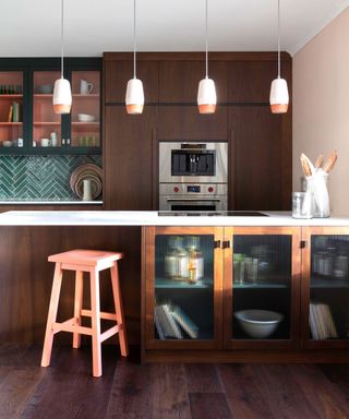 A kitchen with dark brown wooden cabinets, black cabinets with glass fronts and pink shelves, dark green tiiles, and a kitchen island with a white surface, a wooden glass cabinet on it and pendant lights hanging above