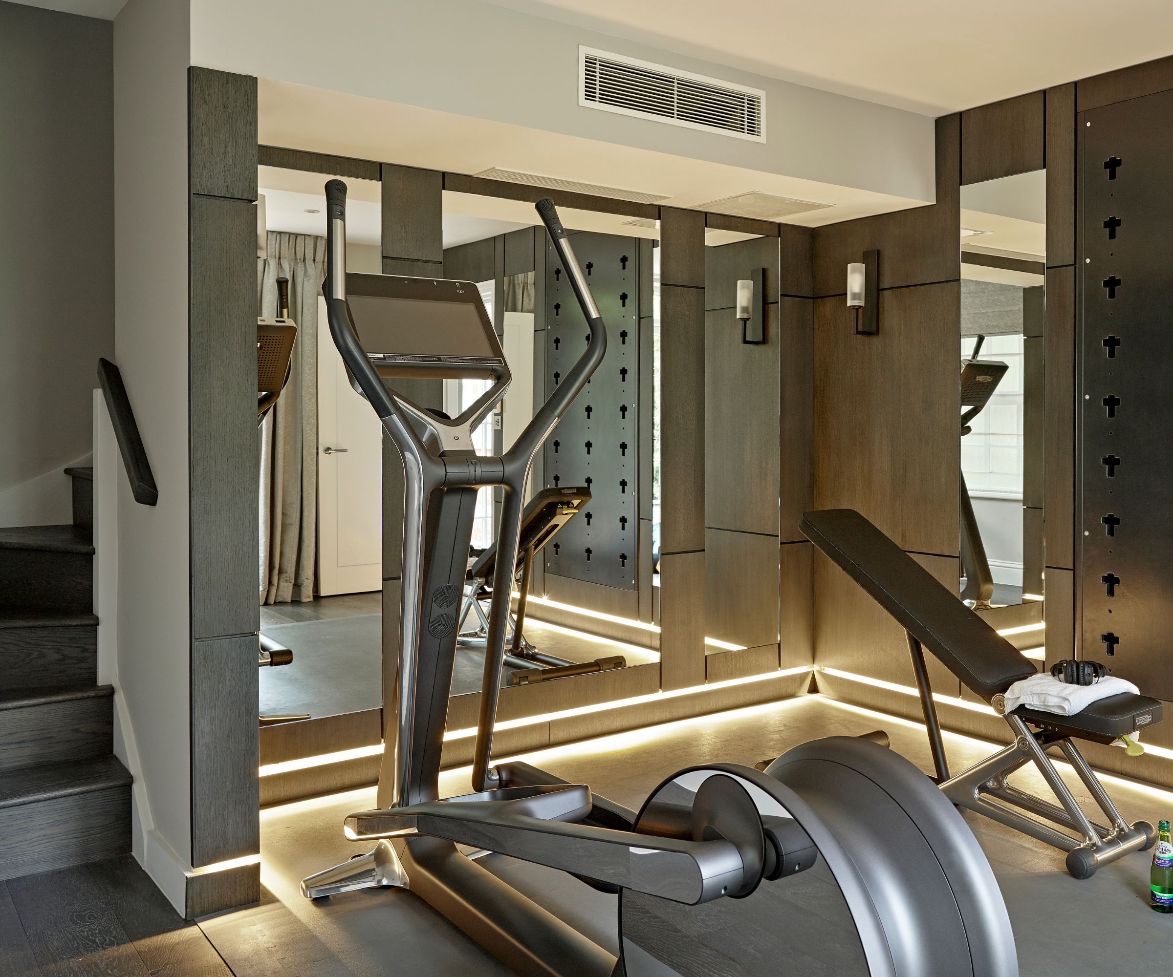 Elliptical trainer and weight bench in dark wood-panelled gym with mirrored walls and strip lighting.