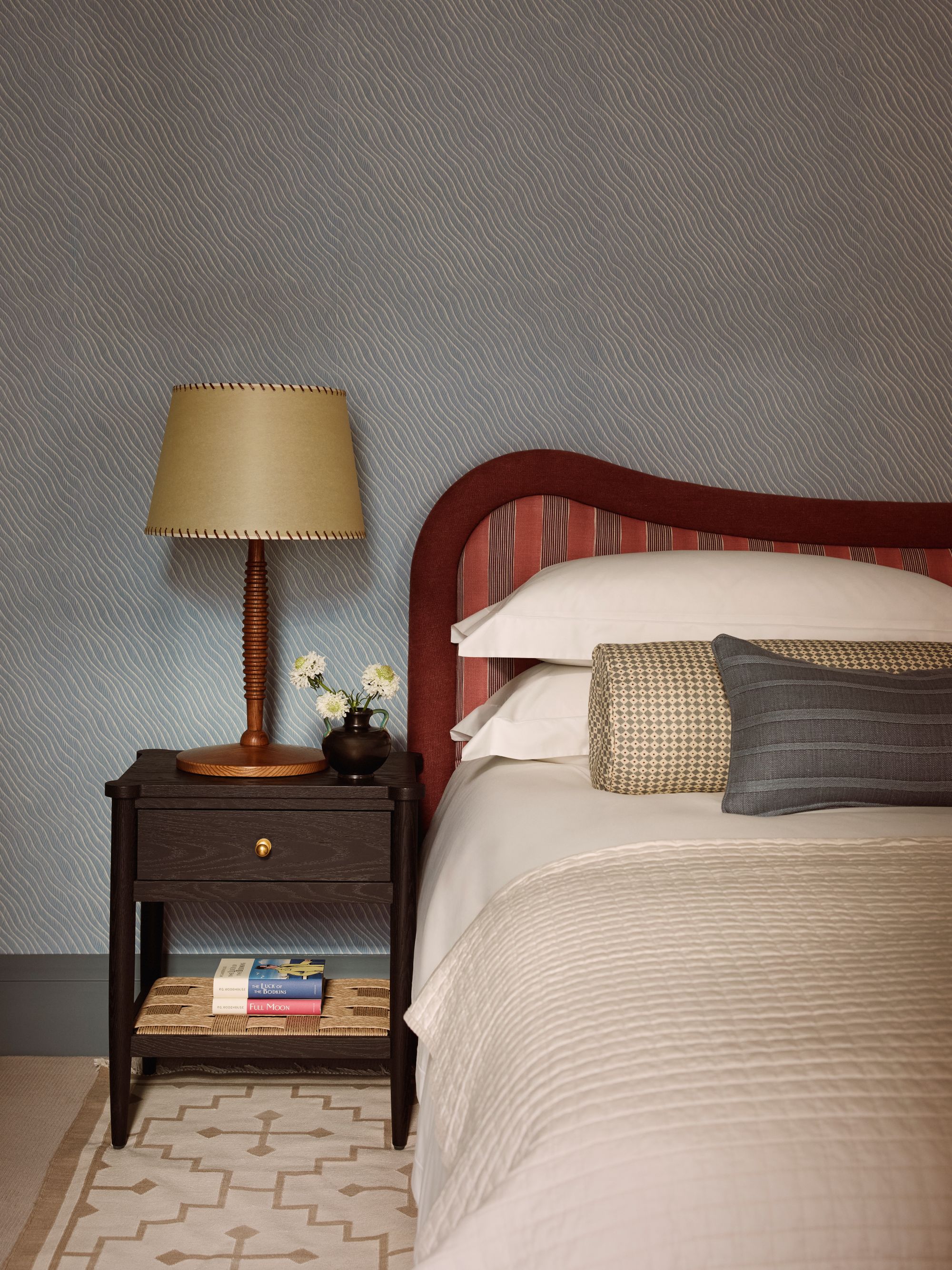 a bedroom with blue and white patterned wallpaper, a tan bedspread, gray rectangular pillow, checkered bolster pillow, red headboard, and dark wood side table