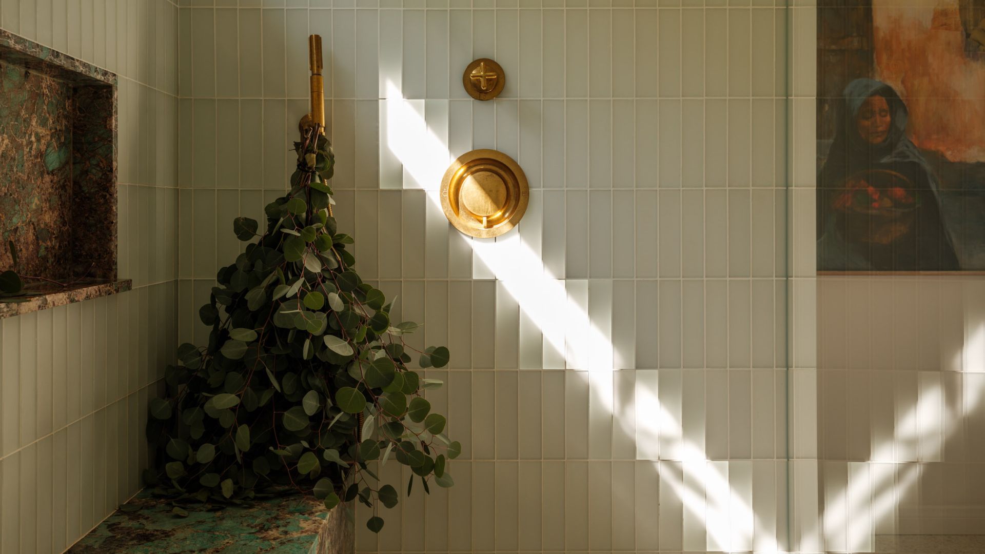 A tiled shower room with a benc. gold hardware, and a branch with eucalyptus